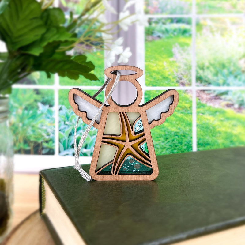 The Forged Flare® Starfish Ornament | 3.5" (H) Mother's Angels® angel figurine stands on a green book by a window, ideal for beach house decor. Greenery and flowers are outside; a vase with green leaves is partly seen on the left.
