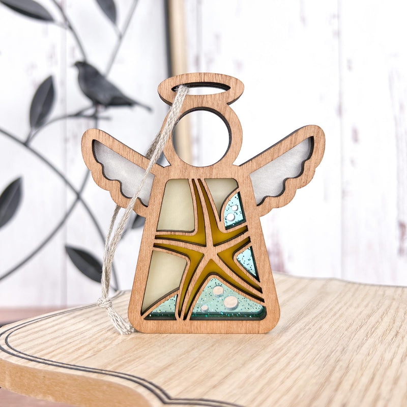 The Forged Flare® Mother's Angels® Starfish Ornament is a 3.5" wooden angel figurine with cutout wings and an abstract blue, yellow, and white design. Includes a string for hanging—perfect beach house decor. Bird-shaped metal accent in background.