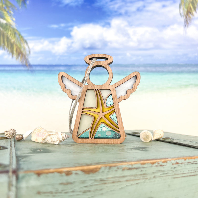 The Forged Flare® Mother's Angels® Starfish Ornament (3.5" H), an angel figurine, stands on a weathered turquoise table with seashells and a tropical beach scene in the background—ideal beach house decor.