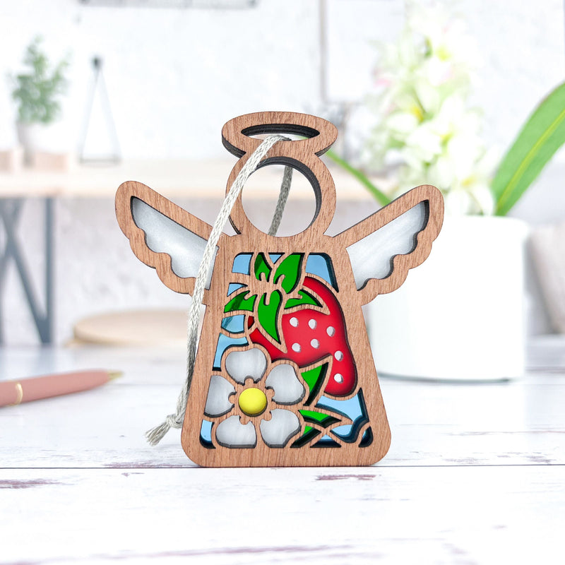 The Forged Flare® Mother's Angels® Strawberry Ornament is a 3.5" tall stained glass-style angel figurine with white flower accents, shown on a white surface—perfect as a charming Christmas tree ornament.