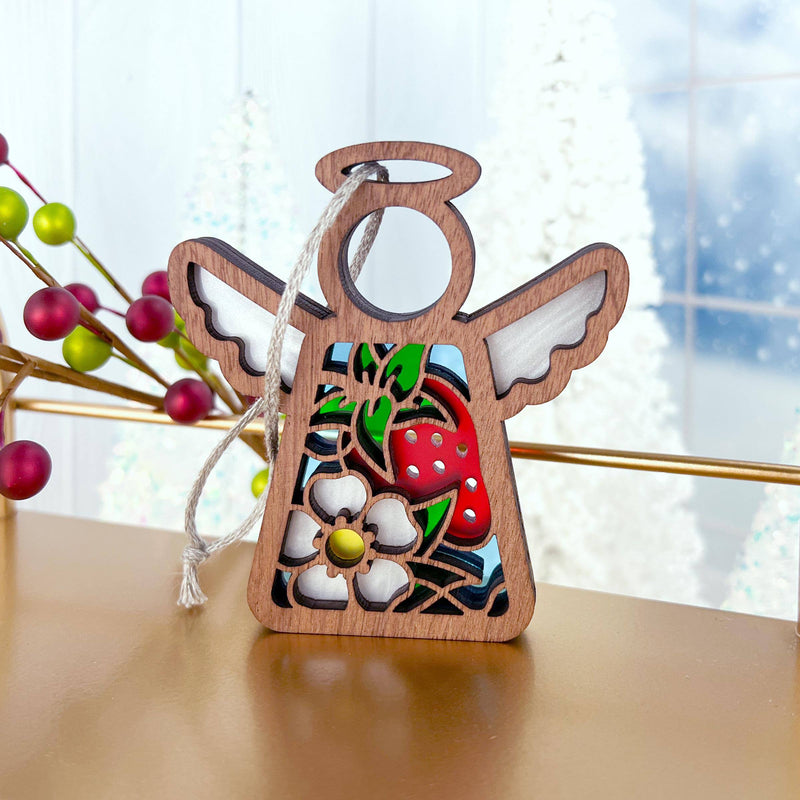 The Forged Flare® Mother's Angels® Strawberry Ornament is a 3.5" wooden angel figurine with cutout designs, a strawberry and white flower, hanging by a silver string amid festive decor of red and green berries and frosted trees.