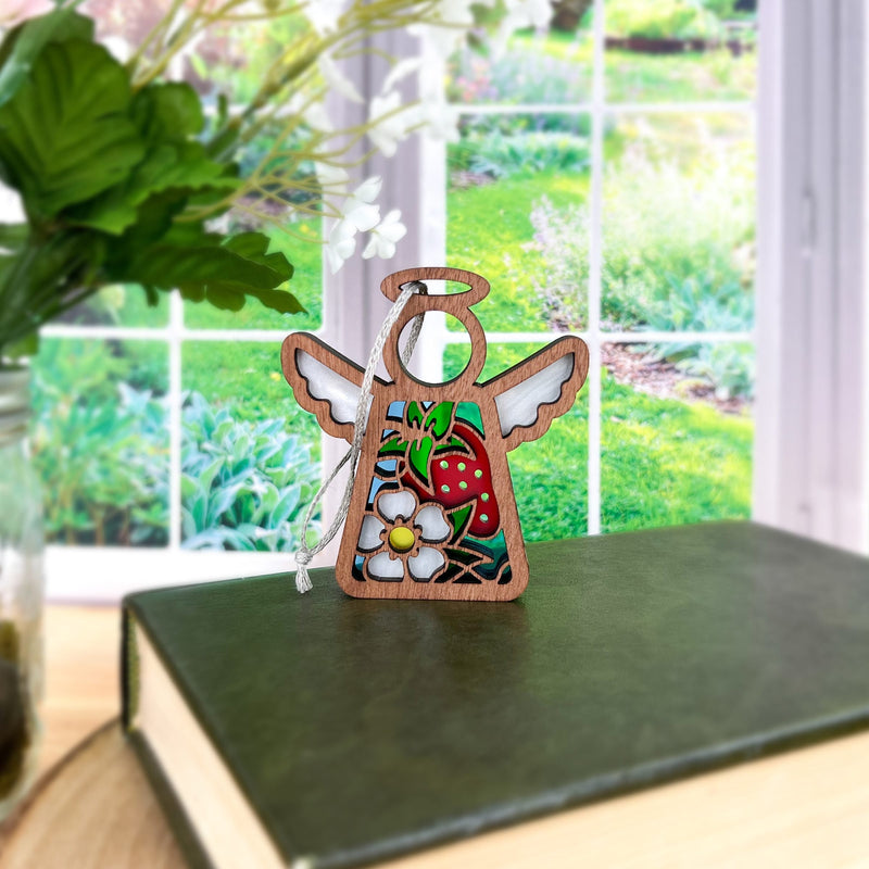 A 3.5" Forged Flare® Mother's Angels® figurine with painted wings, flowers, and a red strawberry ornament stands on a green book by a window, with a leafy plant and garden outside.