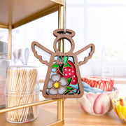 A Forged Flare® Mother's Angels® Strawberry Ornament, a 3.5" wooden angel figurine with white wings and a gray flower, hangs on a gold kitchen shelf near toothpicks and colorful ceramic bowls in a bright kitchen.