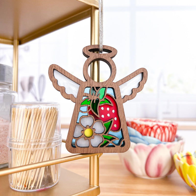 A Forged Flare® Mother's Angels® Strawberry Ornament, a 3.5" wooden angel figurine with white wings and a gray flower, hangs on a gold kitchen shelf near toothpicks and colorful ceramic bowls in a bright kitchen.