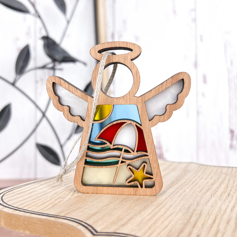 The Forged Flare® Summer Beach Ornament—Mother's Angels® 3.5" angel figurine with stained glass-style beach scene—is a perfect coastal decor piece or unique Christmas tree ornament. Decorative metal bird shown in background.