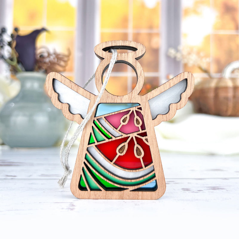The Forged Flare® Summer Watermelon Ornament is a 3.5" Mother's Angels® figurine featuring stained glass-style red, green, and blue sections with white wings, displayed on a white surface with blurred vases and a window behind.
