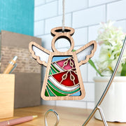 The Summer Watermelon Ornament by Forged Flare® is a 3.5" Mother's Angels® angel figurine with a stained glass-style design in red, green, and white, hanging by a string—ideal for your Christmas tree. A desk, plant, pencils, and tiled wall appear in the background.