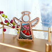 The Forged Flare® Summer Watermelon Ornament features a 3.5" Mother's Angels® wooden angel figurine with stained glass-style accents, hanging on a gold stand amid red berries and frosted white trees in the background.