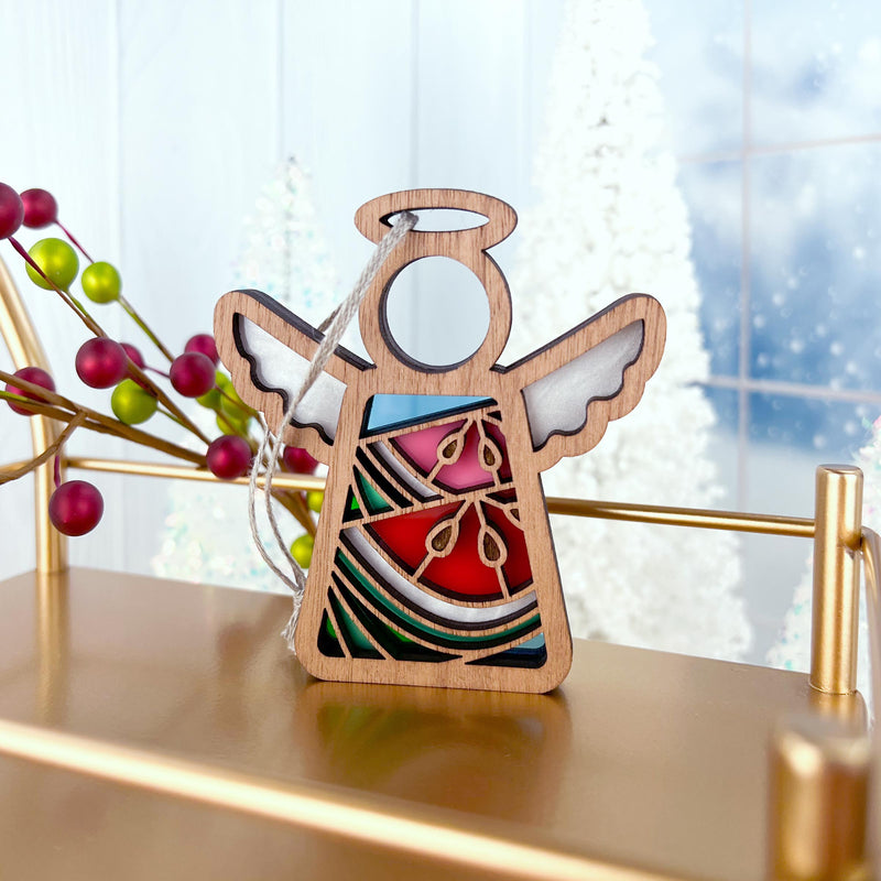 The Forged Flare® Summer Watermelon Ornament features a 3.5" Mother's Angels® wooden angel figurine with stained glass-style accents, hanging on a gold stand amid red berries and frosted white trees in the background.