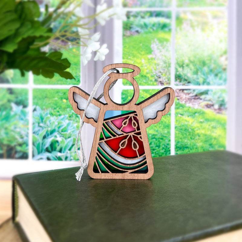 The Forged Flare® Summer Watermelon Ornament is a 3.5" Mother's Angels® angel figurine with colorful stained-glass wings and body, displayed on a green book with a white hanging string. A garden appears beyond large windows in the background.
