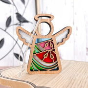 The Summer Watermelon Ornament is a 3.5" angel figurine from Forged Flare® Mother's Angels®, featuring colorful stained glass-style sections and gold accents with a hanging string—ideal as a unique Christmas tree ornament. A bird sculpture sits blurred in the background.
