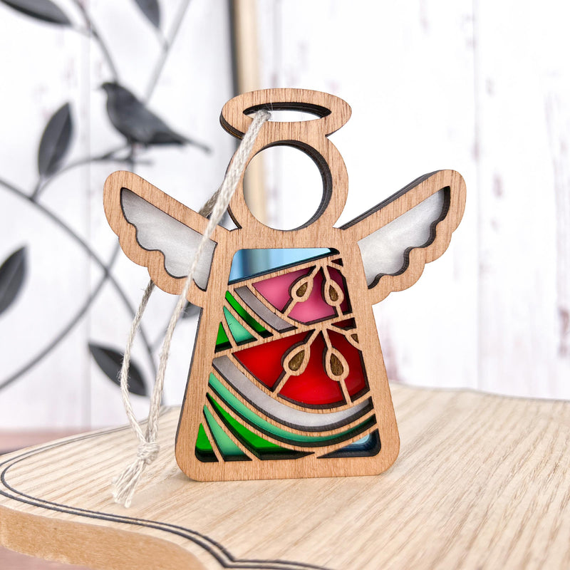 The Summer Watermelon Ornament is a 3.5" angel figurine from Forged Flare® Mother's Angels®, featuring colorful stained glass-style sections and gold accents with a hanging string—ideal as a unique Christmas tree ornament. A bird sculpture sits blurred in the background.