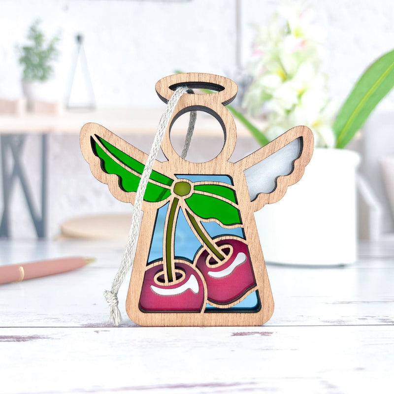 The Sweet Cherries Ornament by Forged Flare® is a 3.5" Mother's Angels® wooden angel figurine with stained glass-style wings and two red cherries, shown on a white surface with blurred plants and decor in the background.