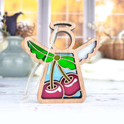 The Sweet Cherries Ornament by Forged Flare® is a 3.5" Mother's Angels® figurine featuring wooden wings and stained glass cherries with green leaves. It has a hanging string and is shown on a white surface with blurred vases and windows behind.