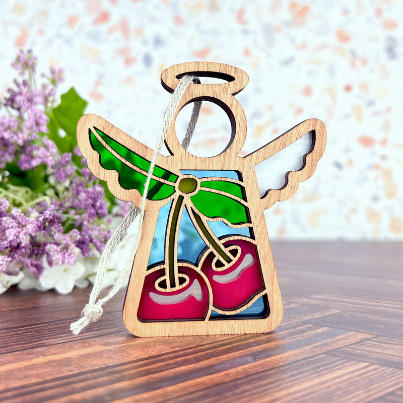 The Forged Flare® Sweet Cherries Ornament is a 3.5" Mother's Angels® figurine with stained-glass-style wings, green ribbon, and red cherries—a charming Christmas accent set off by purple flowers and green leaves in the background.