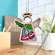 The Sweet Cherries Ornament by Forged Flare® features a 3.5" Mother's Angels® figurine with stained glass-style wings and red cherries, displayed on a silver stand. Flowers, stationery, and a teal wall complete this festive scene.