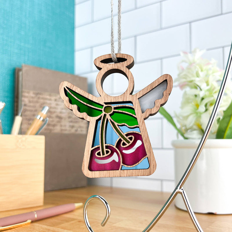 The Sweet Cherries Ornament by Forged Flare® features a 3.5" Mother's Angels® figurine with stained glass-style wings and red cherries, displayed on a silver stand. Flowers, stationery, and a teal wall complete this festive scene.