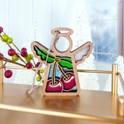 The Forged Flare® Sweet Cherries Ornament, a 3.5" Mother's Angels® wooden angel figurine with painted wings and red cherries, hangs by a string from a gold shelf. Festive decorations and frosted trees blur in the background.