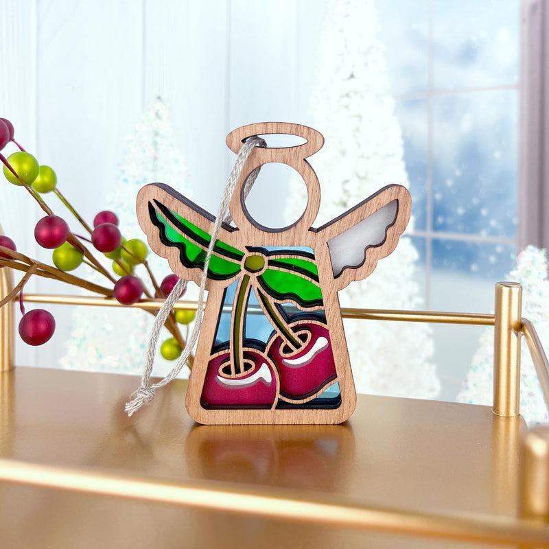 The Forged Flare® Sweet Cherries Ornament, a 3.5" Mother's Angels® wooden angel figurine with painted wings and red cherries, hangs by a string from a gold shelf. Festive decorations and frosted trees blur in the background.