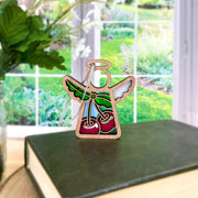 The Sweet Cherries Ornament, a 3.5" (H) angel figurine from Forged Flare's® Mother's Angels®, features vibrant hues and stands on a green book by the window, with plants and a garden visible outside. Perfect as a Christmas ornament.