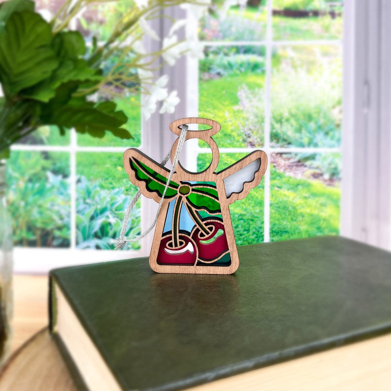 The Sweet Cherries Ornament, a 3.5" (H) angel figurine from Forged Flare's® Mother's Angels®, features vibrant hues and stands on a green book by the window, with plants and a garden visible outside. Perfect as a Christmas ornament.