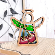 A Forged Flare® Mother's Angels® 3.5" wooden angel figurine, featuring stained glass-style wings and a sweet cherries ornament, hangs by a string on a light wood surface with a blurred bird and branch design in the background.
