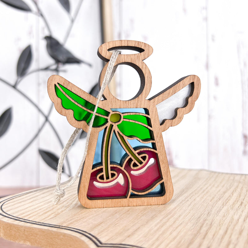 A Forged Flare® Mother's Angels® 3.5" wooden angel figurine, featuring stained glass-style wings and a sweet cherries ornament, hangs by a string on a light wood surface with a blurred bird and branch design in the background.