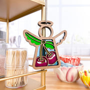 The Forged Flare® Sweet Cherries Ornament, a 3.5" stained glass-style Mother's Angels® angel figurine, hangs on a gold shelf in a bright kitchen beside toothpicks and colorful bowls.