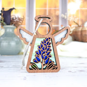 The Forged Flare® Texas Bluebonnet Ornament is a 3.5" Mother's Angels® figurine featuring a stained-glass style bluebonnet design and white wings, displayed on a white surface with blurred vases and autumn flowers in the background.