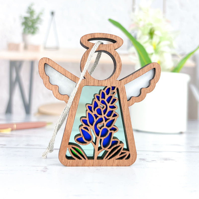 The Forged Flare® Texas Bluebonnet Ornament is a 3.5" Mother's Angels® wooden angel figurine with white wings and bluebonnet decor, shown on a white surface with blurred plants and objects in the background.
