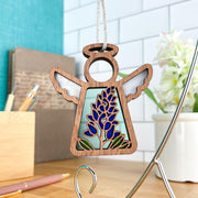 The Forged Flare® Texas Bluebonnet Ornament | 3.5" (H) Angel Figurine from the Mother's Angels® collection, with stained glass wings and a wooden design, hangs on a silver string in a modern room with floral décor and a potted plant.