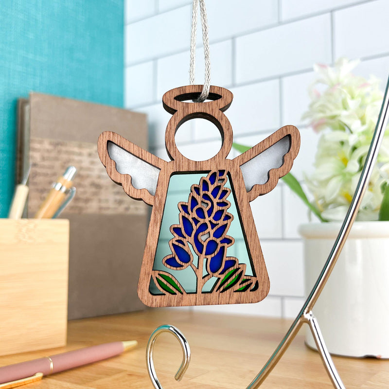 The Forged Flare® Texas Bluebonnet Ornament | 3.5" (H) Angel Figurine from the Mother's Angels® collection, with stained glass wings and a wooden design, hangs on a silver string in a modern room with floral décor and a potted plant.