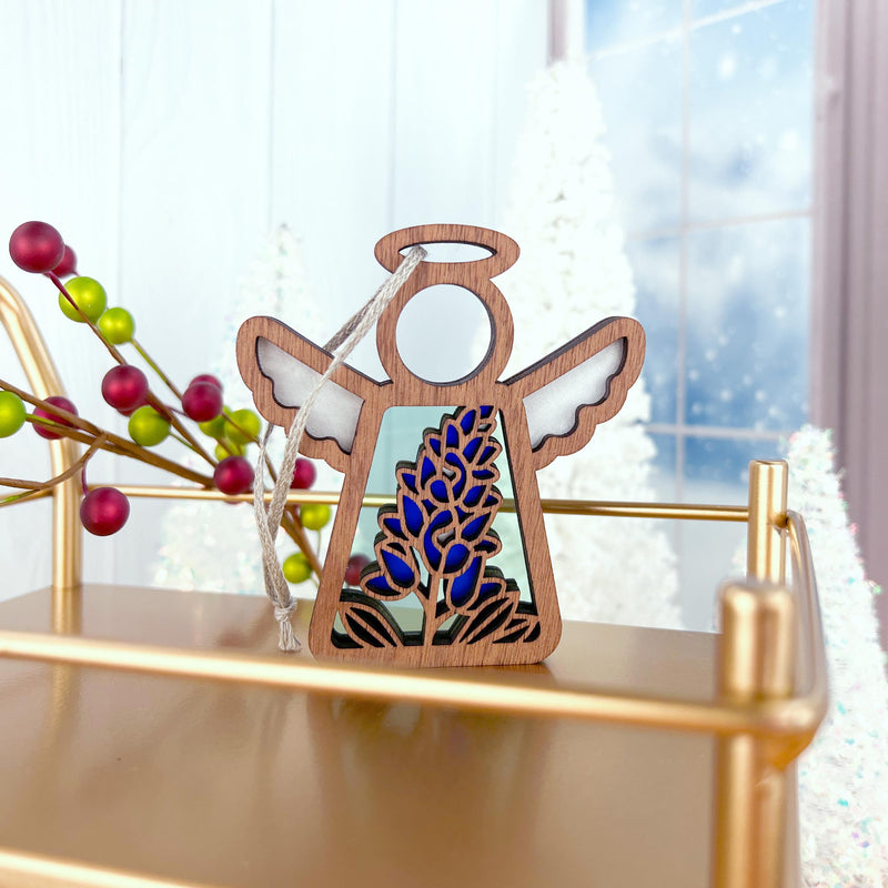 The Texas Bluebonnet Ornament by Forged Flare® is a 3.5" Mother's Angels® figurine, displayed on a gold tray amid festive berries and frosted trees—a lovely flower decor piece in a bright, wintry scene.