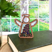 The Forged Flare® Texas Bluebonnet Ornament, a 3.5" Mother's Angels® figurine with white wings and blue and purple floral design, stands on a green book with a sunlit garden visible through a large window in the background.