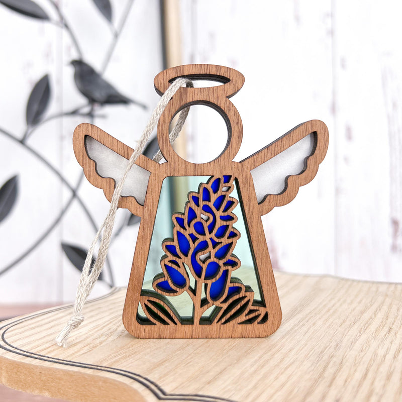 The Forged Flare® Texas Bluebonnet Ornament is a 3.5" Mother's Angels® figurine featuring a stained glass bluebonnet, white wings, and a hanging string—shown on light wood with a blurred plant for lovely floral decor.