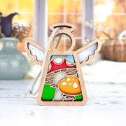 The Forged Flare® Mother's Angels® Toadstool Mushroom Ornament is a 3.5" wooden angel figurine with cutout wings and a halo, accented in red and orange. It stands on a white surface with blurred vases and autumn decor in the background.