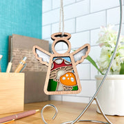 The Forged Flare® Mother's Angels® Toadstool Mushroom Ornament is a 3.5" wooden angel figurine with stained glass and vibrant red-orange mushroom accents, hanging from a string in front of pens, books, and a white tiled wall.
