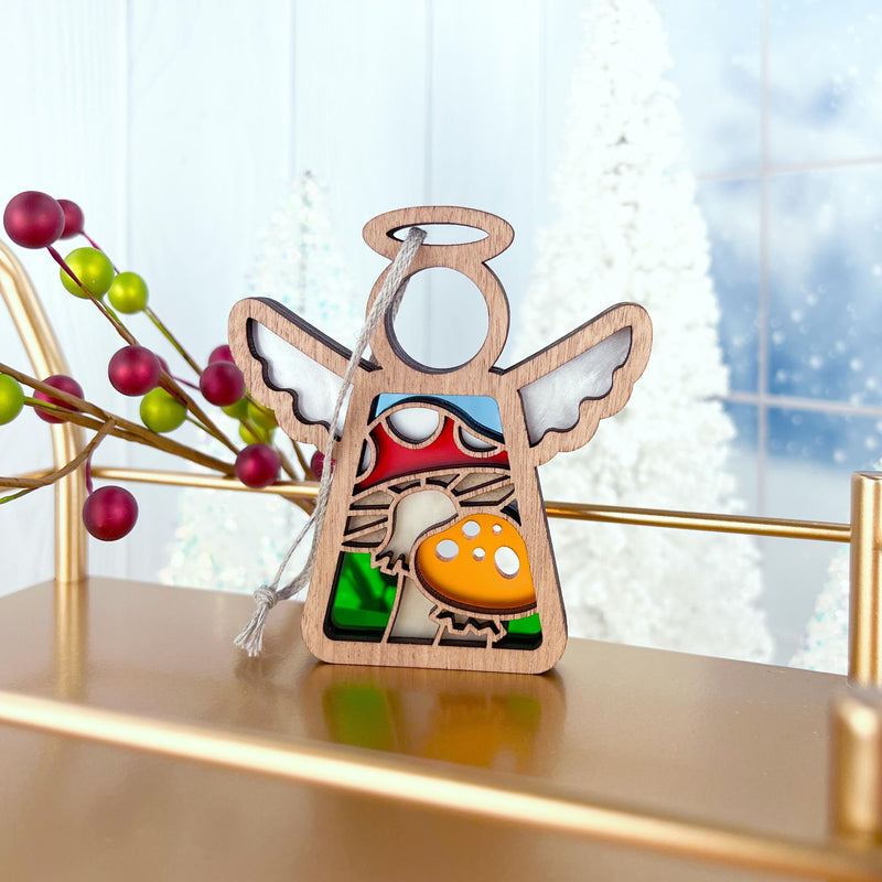 A Forged Flare® Mother's Angels® 3.5" angel figurine with stained glass wings and a colorful toadstool mushroom ornament stands on a gold shelf, framed by red berries, white Christmas trees, and a frosty window.