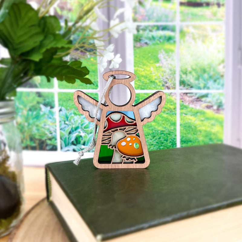 The Forged Flare® Mother's Angels® 3.5" wooden angel figurine features a stained glass-style toadstool mushroom ornament and sits on a green book near a sunlit window, with greenery, flowers, and a jar of white blooms nearby.