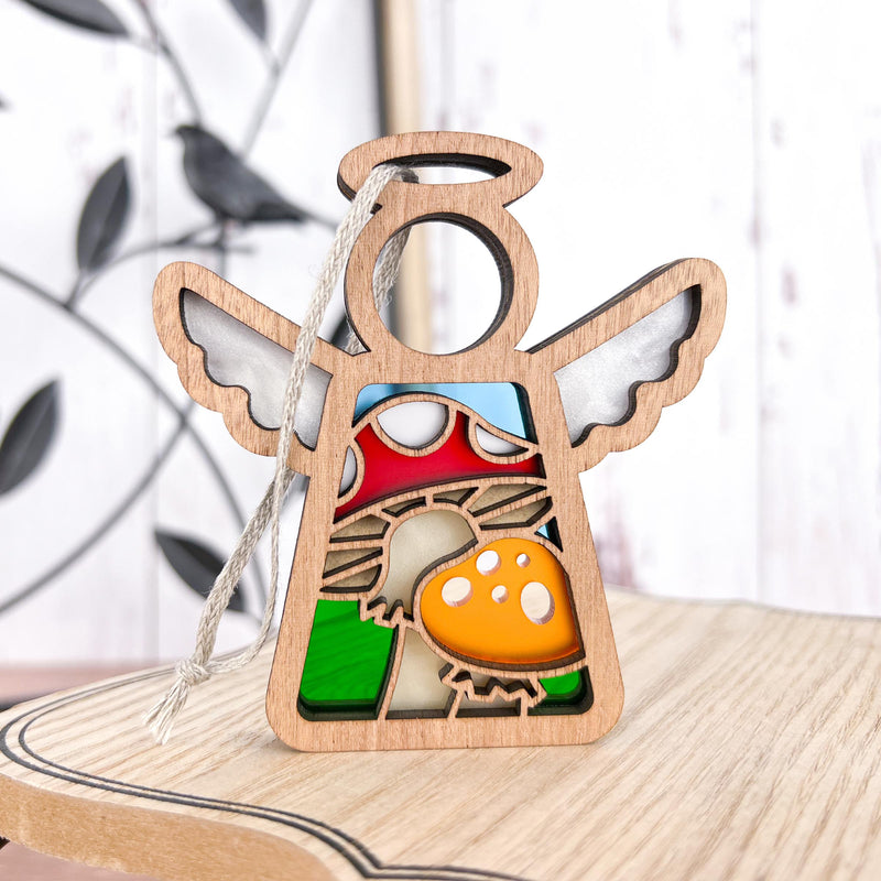 The Forged Flare® Mother's Angels® Toadstool Mushroom Ornament is a 3.5" wooden angel figurine with cut-out wings, featuring stained glass-style red and yellow mushrooms and green grass—perfect as a charming Christmas tree decoration.