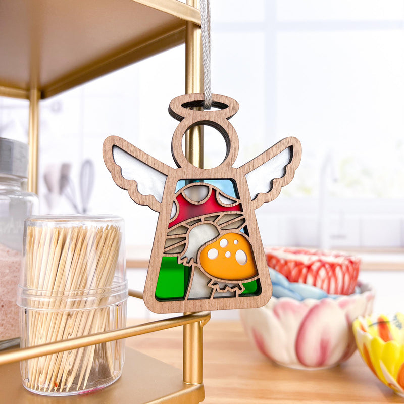 The Forged Flare® Toadstool Mushroom Ornament | 3.5" (H) Angel Figurine from the Mother's Angels® collection hangs on a kitchen shelf beside toothpicks and pastel ceramic bowls, its stained glass accents adding charm.