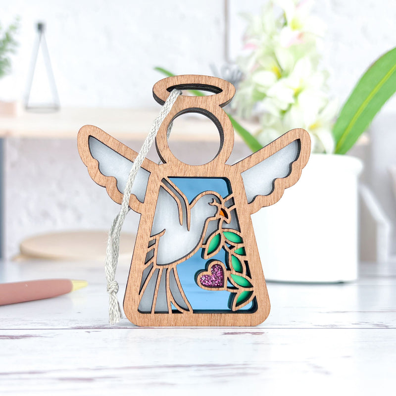 The Forged Flare® Mother's Angels® 3.5" angel figurine, featuring a white dove with an olive branch and a pink heart, stands on a table—a thoughtful memorial gift.