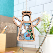 The Forged Flare® White Dove Ornament | 3.5" (H) Angel Figurine by Mother's Angels® features a cutout angel and purple heart, hanging by a string—an ideal memorial or bird lover gift. Flowers, pen, and tiled wall are softly visible in the background.