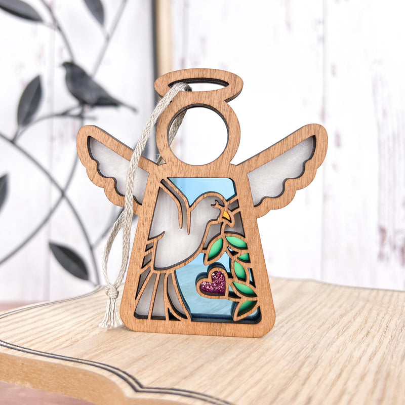 The Forged Flare® Mother's Angels® White Dove Ornament is a 3.5" wooden angel figurine with a glittery pink heart and olive branch, perfect as a memorial or bird lover gift. Hangs by a cord on light wood; bird and branch decor in background.