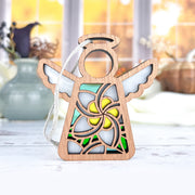 The Forged Flare® White Plumeria Ornament is a 3.5" Mother's Angels® figurine with a hollow center, yellow plumeria flower, green accents, white wings, and jute string—resting on a white surface with a blurred indoor background.