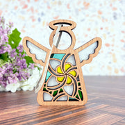 The Forged Flare® White Plumeria Ornament is a 3.5" angel figurine with intricate cut-out designs and yellow-green accents, displayed on wood with purple and white plumeria-like flowers in the background.