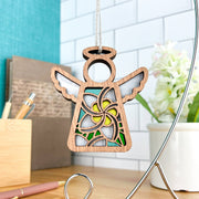 The Forged Flare® White Plumeria Ornament is a 3.5" Mother's Angels® wooden angel with floral cutouts, hanging on a string and displayed on a desk with pens, books, and a potted plant against a tile wall.