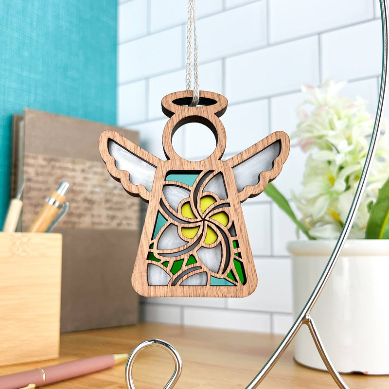 The Forged Flare® White Plumeria Ornament is a 3.5" Mother's Angels® wooden angel with floral cutouts, hanging on a string and displayed on a desk with pens, books, and a potted plant against a tile wall.