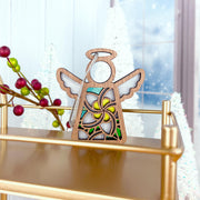 The Forged Flare® White Plumeria Ornament is a 3.5" Mother's Angels® angel figurine with a floral design, displayed on a gold shelf with red berries and white Christmas trees by a frosted window for a festive Hawaiian touch.