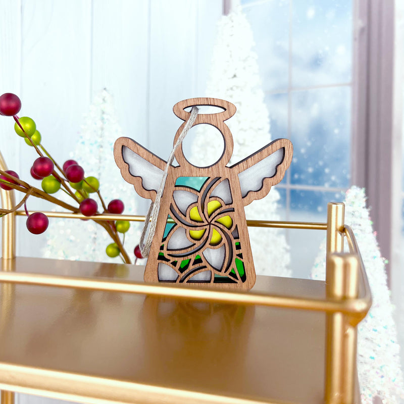 The Forged Flare® White Plumeria Ornament is a 3.5" Mother's Angels® angel figurine with a floral design, displayed on a gold shelf with red berries and white Christmas trees by a frosted window for a festive Hawaiian touch.